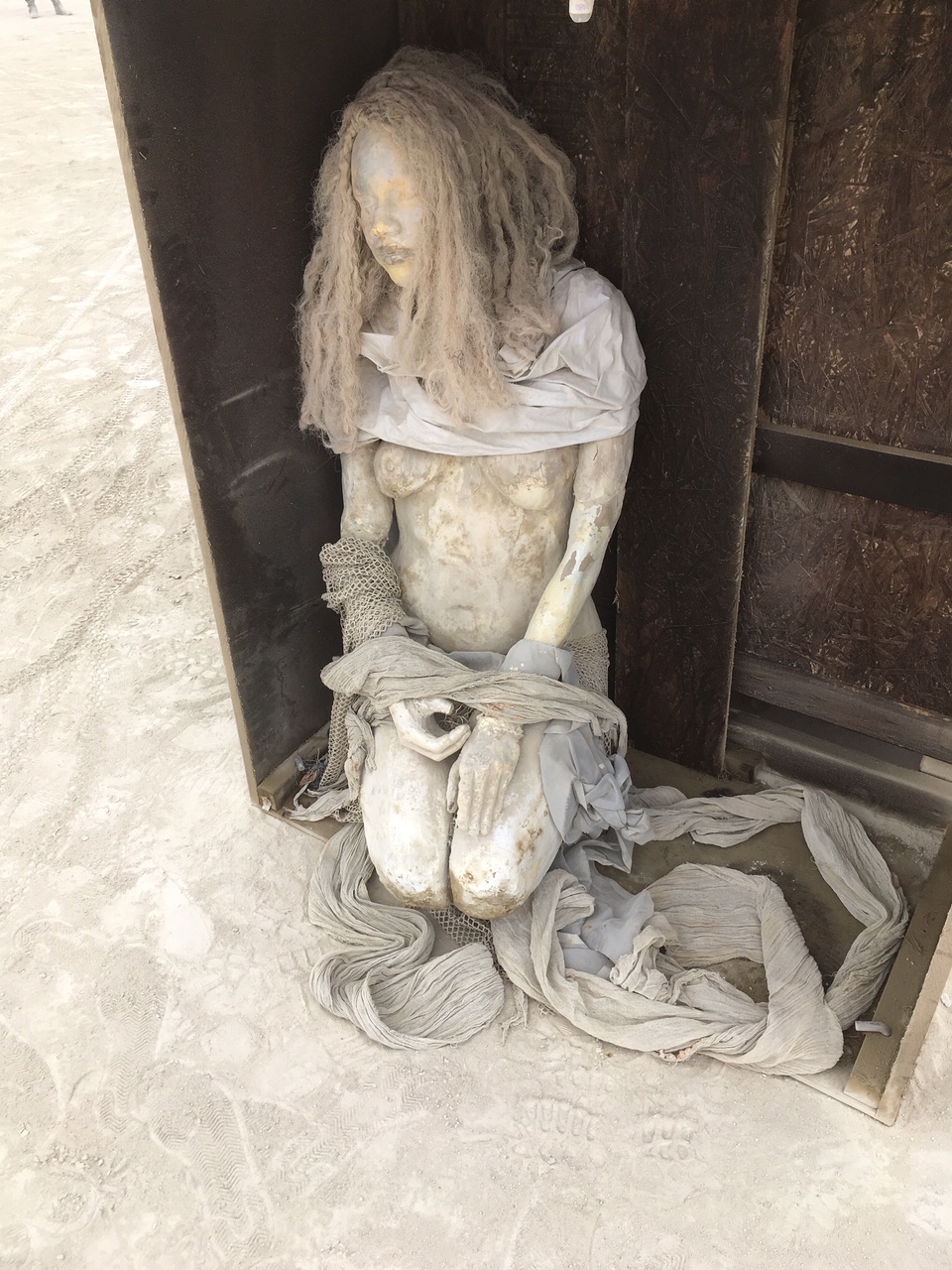 Kneeling cast figure with long hair inside a weathered wooden box on the playa