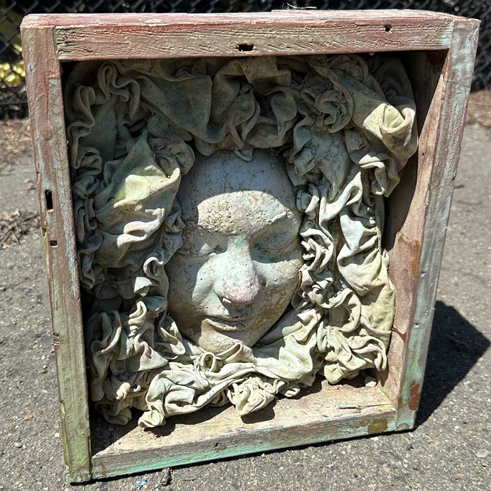 Close view of a face cast in a weathered wooden box, framed by ruffled, sculpted fabric