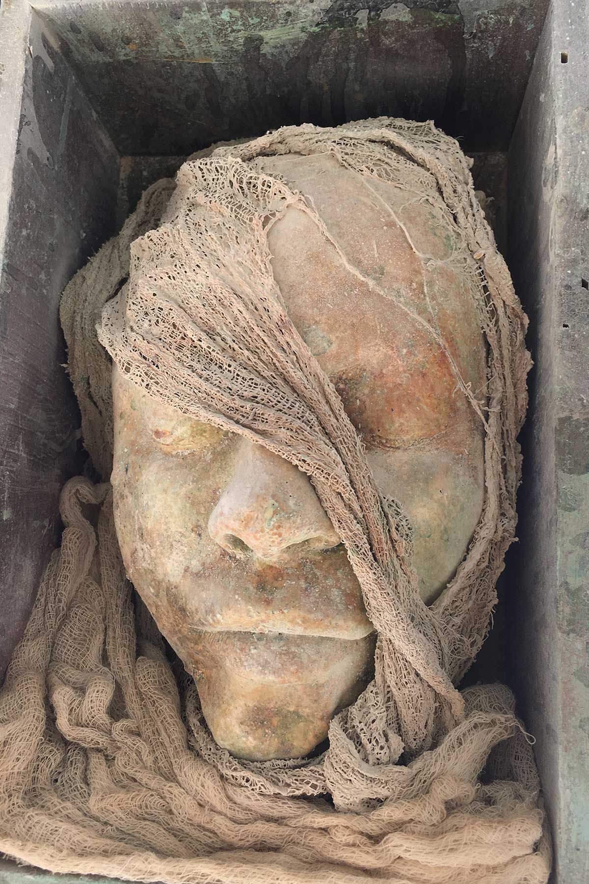 Face cast in a wooden box, partly veiled by fabric, with a warm rust-toned patina