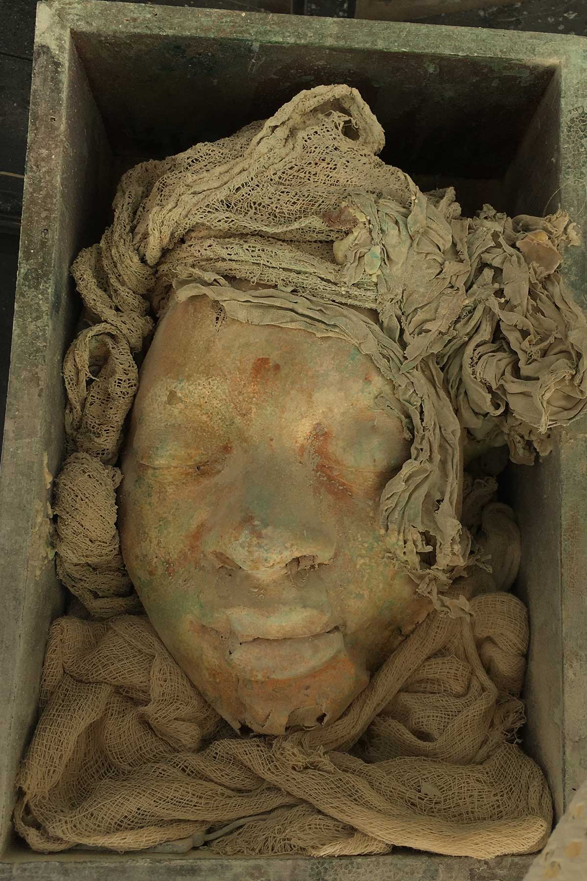 Smiling face cast in a wooden box, surrounded by sculpted fabric with a rust-toned patina