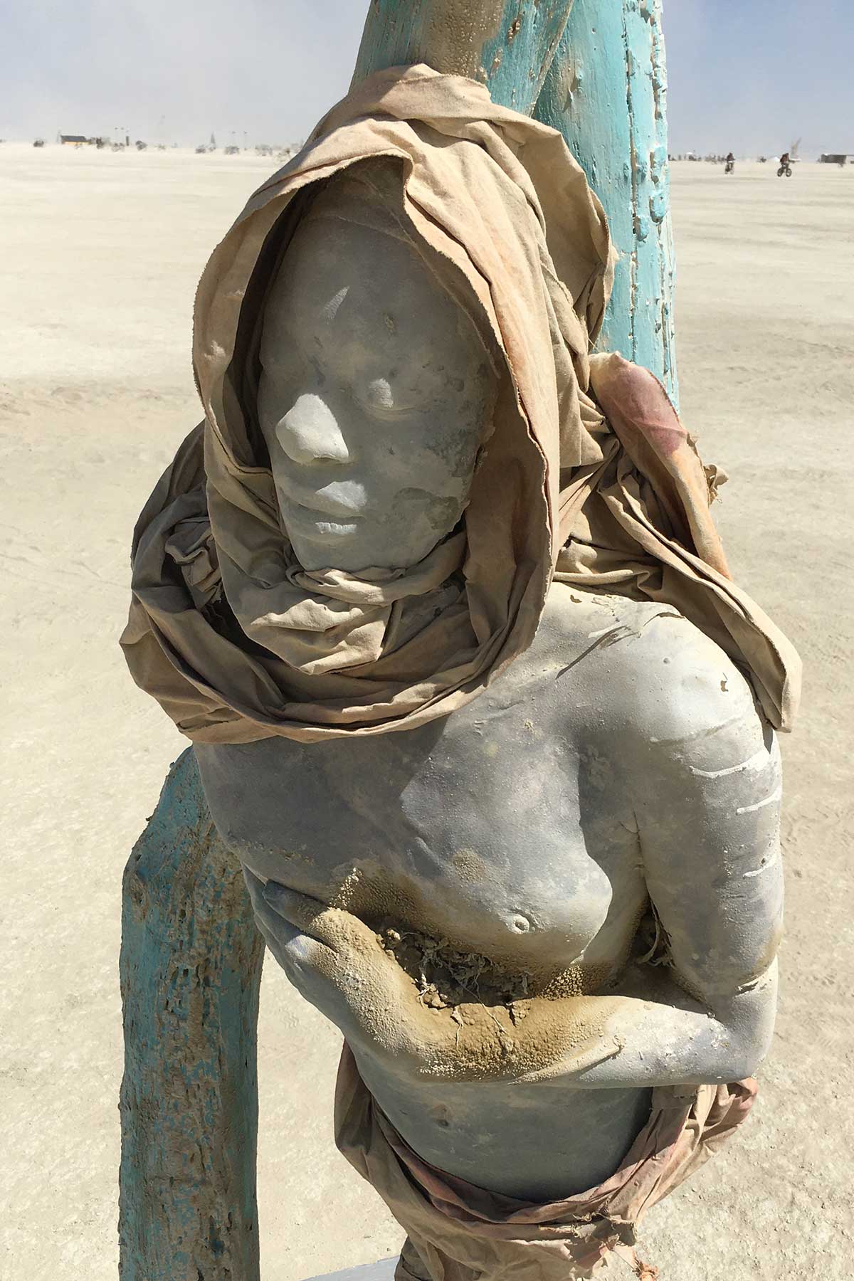 Hooded cast figure leaning against a teal post on the playa