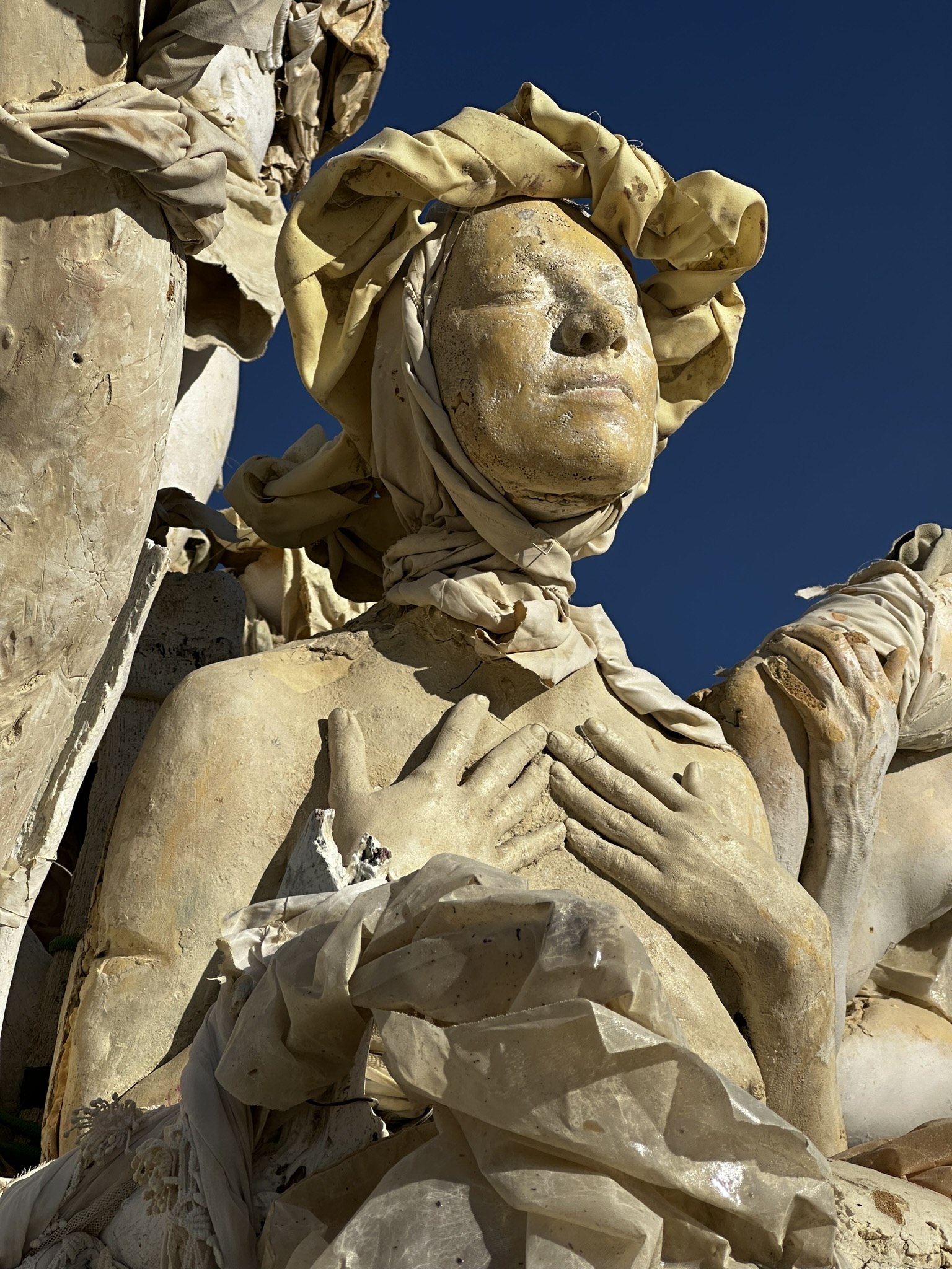 Cast figure with a sculpted head wrap and hand over heart, against a deep blue sky