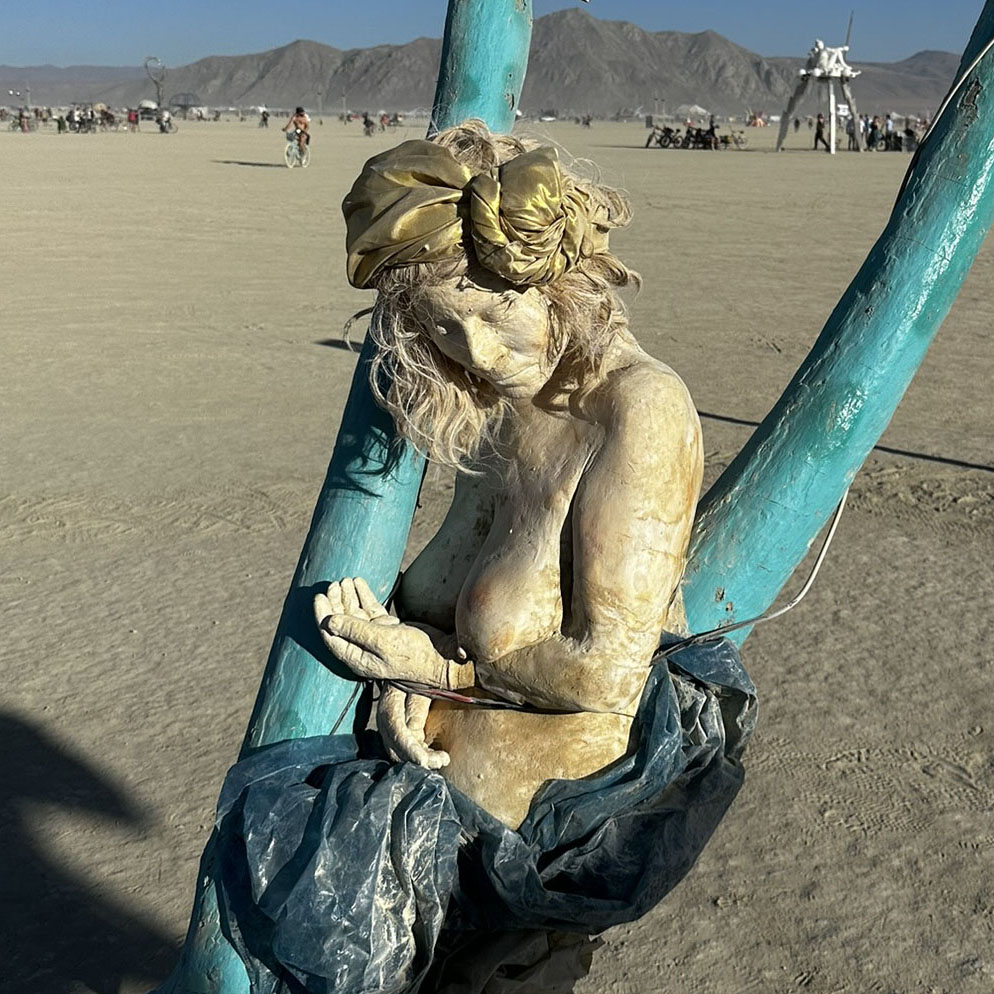 Cast figure with a head wrap and blue fabric, cradled between teal poles on the playa